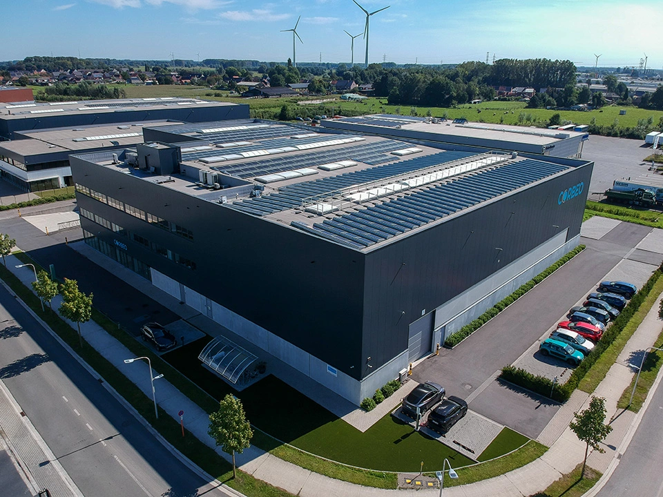 Aerial view of a modern industrial building with the logo “CORBEO” on the side, surrounded by parking lots, green spaces, and wind turbines in the background.