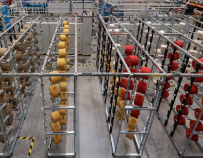 Industrial setup with rows of metal racks holding large spools of thread or yarn in beige, yellow, and red colors.